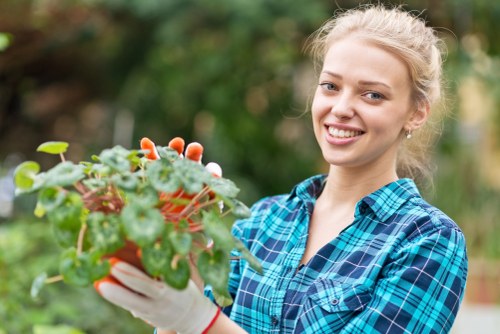 A young woman with blonde hair tied back, wearing a blue and black checkered shirt and gardening gloves, is standing outdoors in a garden. She is holding a small potted plant with green leaves and pinkish-red flowers, smiling at the camera. The garden background shows a blurred view of greenery, including trees and shrubs, suggesting a well-maintained outdoor space suitable for gardening services in Sudbury. The natural light indicates a bright, clear day, highlighting the healthy plant and the vibrant appearance of the garden environment, which may include lawn areas, flower beds, and garden borders. This scene reflects typical tasks involved in professional gardening and landscaping work, such as planting and maintaining lush, colorful gardens in the local area.
